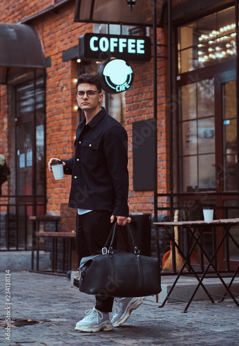 A handsome man in glasses wearing a shirt holding a takeaway coffee and bag while walking down the street.