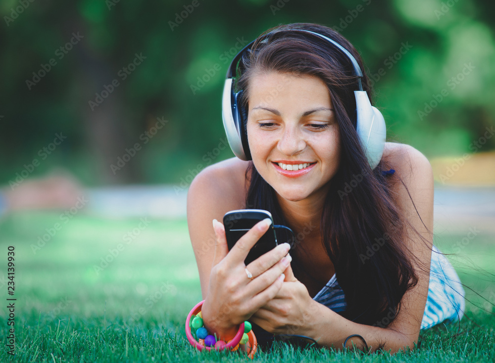 Beautiful young woman with headphones lying down on green lawn in a park. Spring time season time Beautiful young woman with headphones lying down on green lawn in a park. Spring time season time