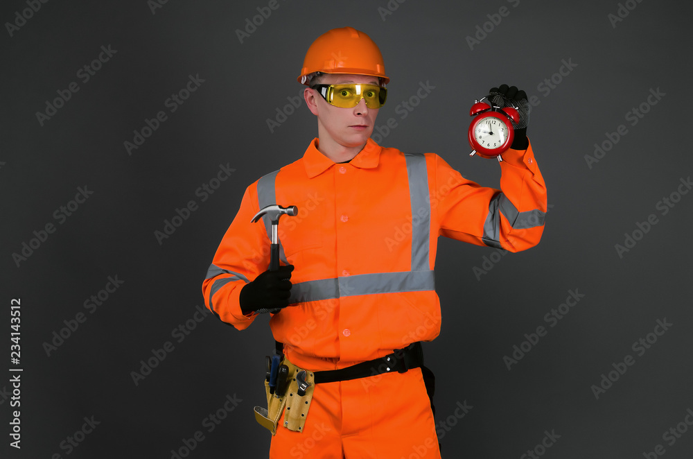Builder worker holding in hand a red alarm clock isolated on gray background. Time to work. Building construction time. Builder worker holding in hand a red alarm clock isolated on gray background. Time to work. Building construction time.