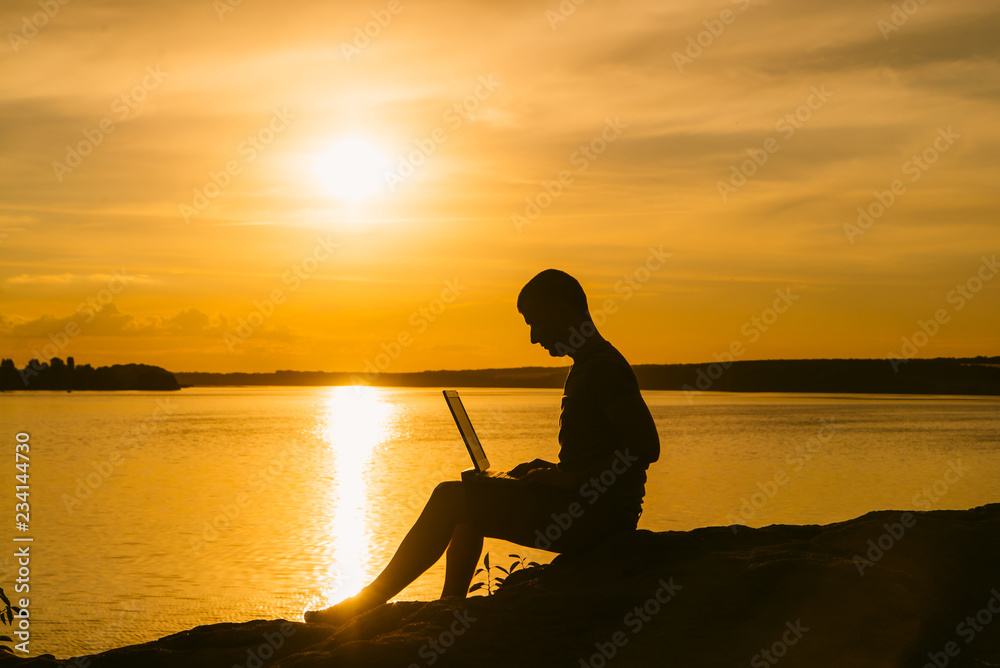 Busy man working on his laptop near the calm evening river during sunset. Silhouette of businessman working with laptop on the sunset background.
