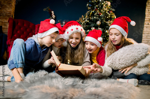 Charming and beautiful little children in Santa's hats are reading Christmas stories and laughing, on the background of a Christmas tree.