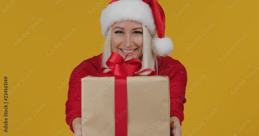 Happy excited young blond woman in Santa Claus red hat gives a gift in a box with a red ribbon over yellow background. Wow surprise effect Happy excited young blond woman in Santa Claus red hat gives a gift in a box with a red ribbon over yellow background. Wow surprise effect