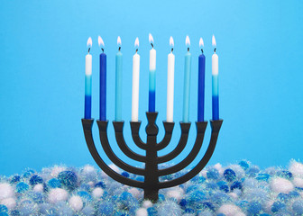 a nine-branched menorah (also called a Chanukiah or Hanukiah) burning candles to celebrate Hanukkah against a light blue background with light and dark blue and white shiny yarn balls underneath.
