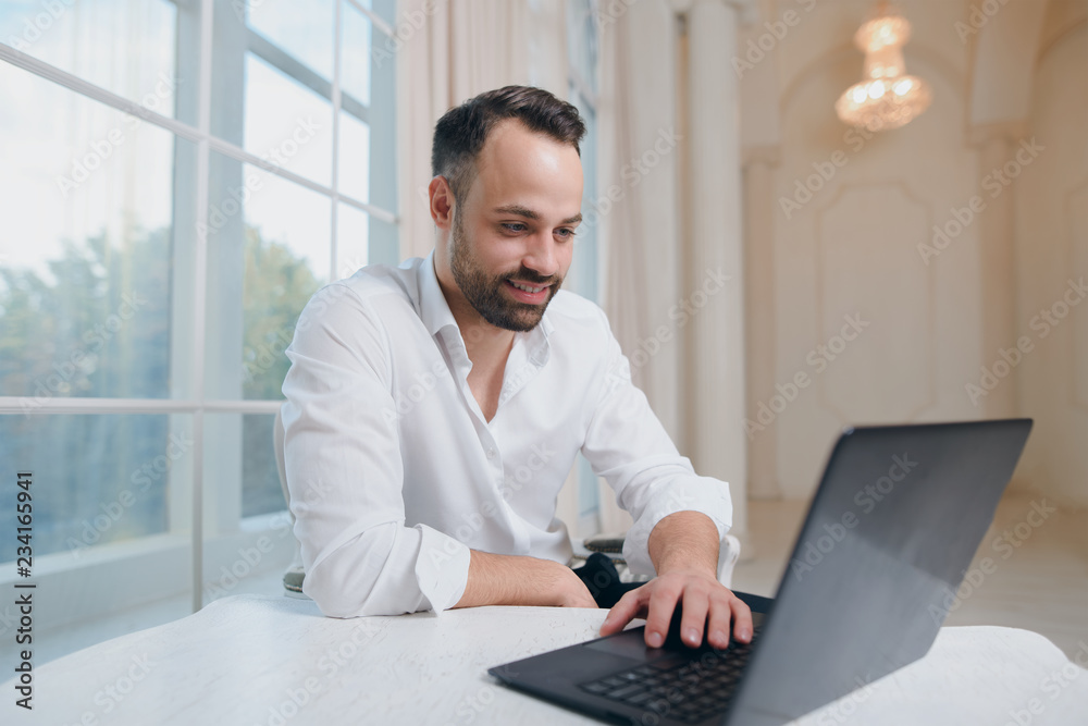 Smiling businessman typing at laptop on window background Smiling businessman typing at laptop on window background