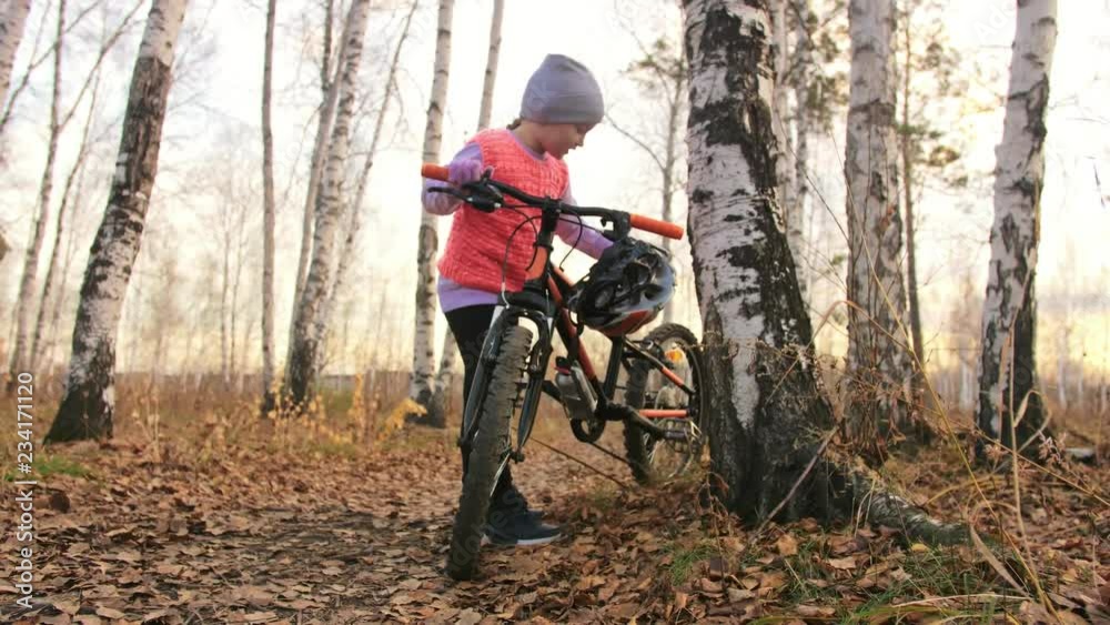 One caucasian children walk with bike in autumn park. Little girl walking black orange cycle in forest. Kid goes do bicycle sports. Biker motion ride with backpack and helmet. Mountain bike hardtail. One caucasian children walk with bike in autumn park. Little girl walking black orange cycle in forest. Kid goes do bicycle sports. Biker motion ride with backpack and helmet. Mountain bike hardtail.