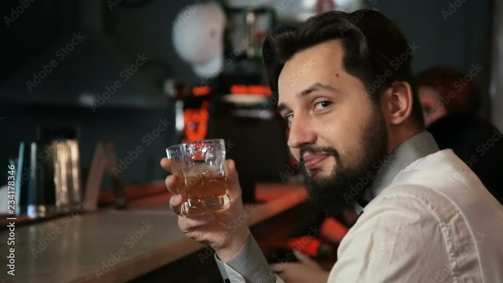 A brunette man sits alone in a bar and drinks whiskey. Smiles, happy. A brunette man sits alone in a bar and drinks whiskey. Smiles, happy.