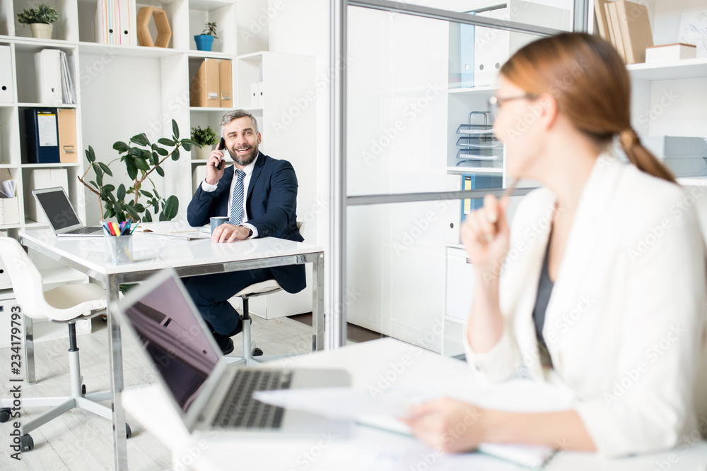 Positive confident handsome male manager in formalwear sitting at table and talking by mobile phone while conferring with colleague in office Positive confident handsome male manager in formalwear sitting at table and talking by mobile phone while conferring with colleague in office