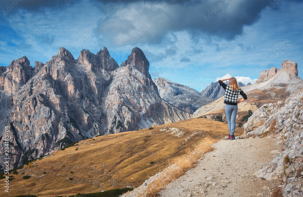 Young woman on the trail is looking on majestic mountains at sunset in autumn in Dolomites, Italy. Landscape with girl, cloudy sky, orange grass, high rocks and trees in fall. Travel in italian alps