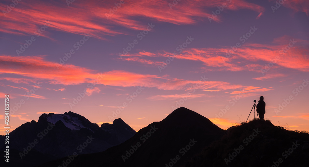 Silhouette of a photographer on the mountain peak against rocks and purple sky with pink clouds at sunset. Landscape with man, mountain ridge and colorful sky at dusk in Dolomites, Italy. Travel
