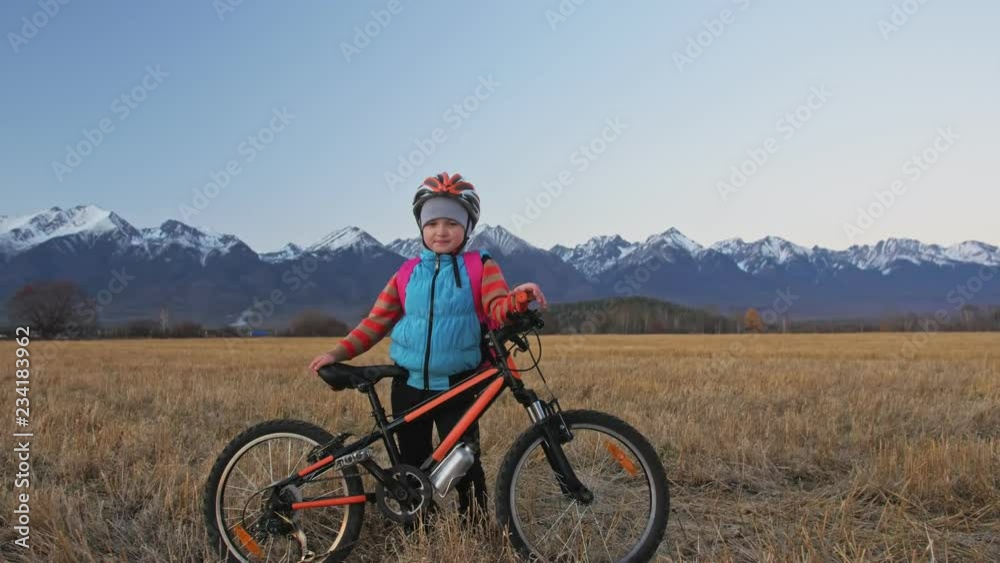 One caucasian children walk with bike in wheat field. Girl walking black orange cycle on background of beautiful snowy mountains. Biker motion ride with backpack and helmet. Mountain bike hardtail. One caucasian children walk with bike in wheat field. Girl walking black orange cycle on background of beautiful snowy mountains. Biker motion ride with backpack and helmet. Mountain bike hardtail.