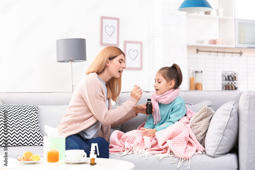Mother giving daughter cough syrup on sofa at home