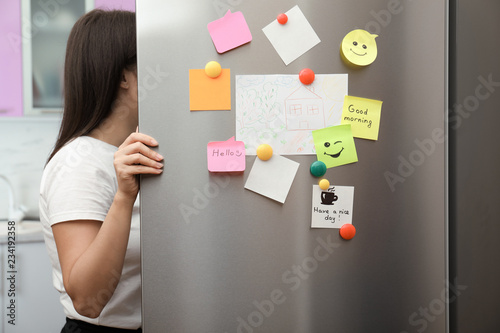 Woman opening refrigerator door with paper sheets and magnets at home, closeup