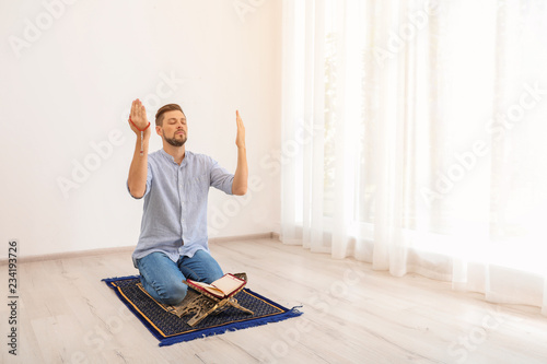 Muslim man with Koran praying on rug indoors. Space for text
