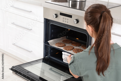Young woman taking baking sheet with cookies from oven at home