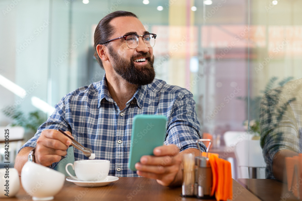 Some sugar. Bearded smiling man putting some sugar into his coffee sitting in cafeteria holding his phone Some sugar. Bearded smiling man putting some sugar into his coffee sitting in cafeteria holding his phone