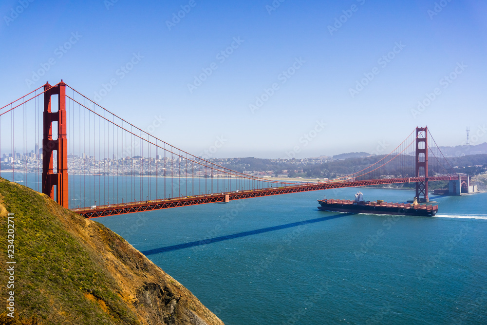 Cargo ship passing under Golden Gate Bridge on a sunny day; San Francisco skyline in the background; California