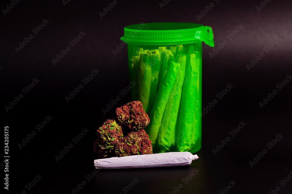 A bright transparent green RX pill bottle filled with marijuana joints next to a pile of buds with long red hairs and a joint laying in front.