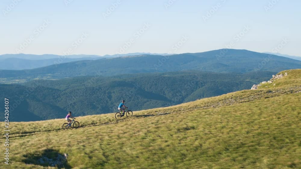 AERIAL: Active young couple riding their mountain bikes along a scenic path running down the grassy hill. Female and male tourist get off their bicycles to look at the picturesque nature below them. AERIAL: Active young couple riding their mountain bikes along a scenic path running down the grassy hill. Female and male tourist get off their bicycles to look at the picturesque nature below them.