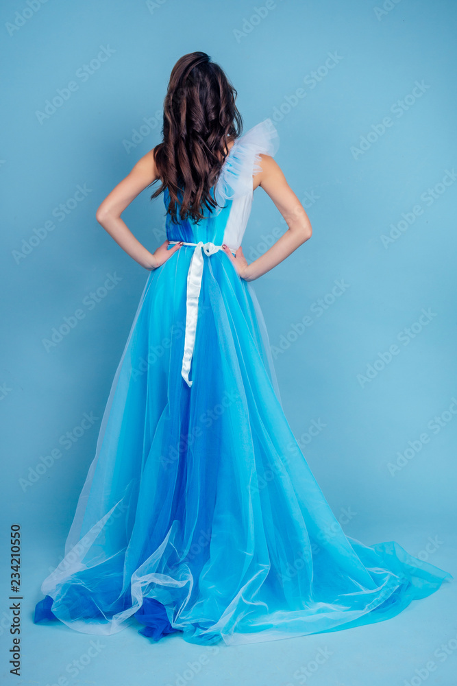 Young beauty woman in tulle fluttering blue dress.Beautiful girl with make-up , hairstyle and gorgeous look.female model prom seamstress and designer clothes tailor on a blue background in the studio