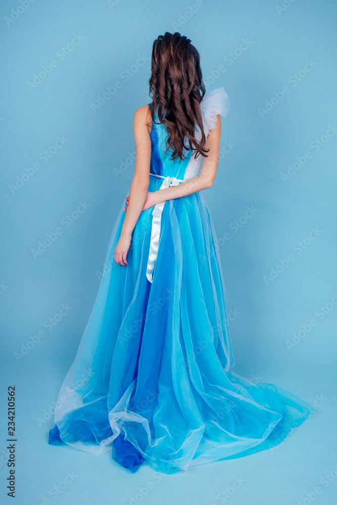 Fototapeta premium Young beauty woman in tulle fluttering blue dress.Beautiful girl with make-up , hairstyle and gorgeous look.female model prom seamstress and designer clothes tailor on a blue background in the studio