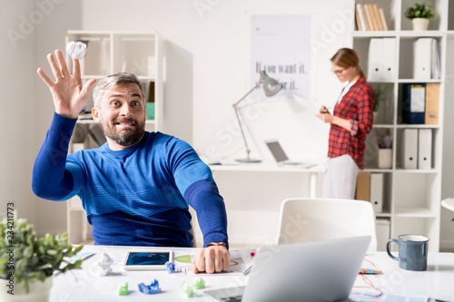Positive playful handsome bearded programmer in sweater sitting at table and throwing paper ball while wasting time in modern office