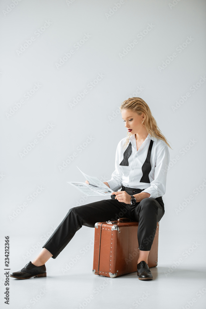 fashionable girl reading newspaper while sitting on retro suitcase on grey fashionable girl reading newspaper while sitting on retro suitcase on grey