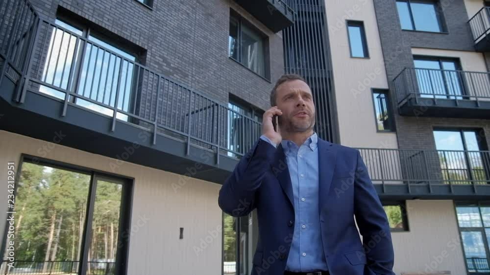 Smiling man standing at residential luxury backyard and speaking at smartphone slow motion. Caucasian ambitious manager in suit wear selling living real estate at mobile speach outroor. Success leader Smiling man standing at residential luxury backyard and speaking at smartphone slow motion. Caucasian ambitious manager in suit wear selling living real estate at mobile speach outroor. Success leader