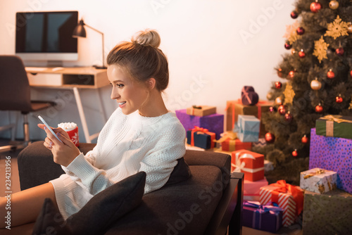 smiling young blonde woman sitting on couch and using smartphone at christmas time