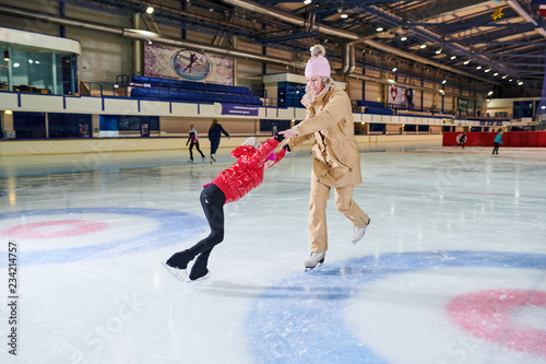 Full length portrait of little girl learning to figure skate with mother in indoor skating  rink, copy space