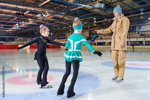 Full  length portrait of woman coach  training  two little girls figure skating in indoor rink