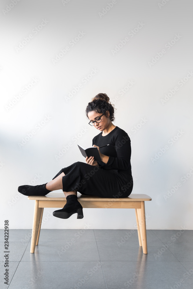 Beautiful woman with curly hair dressed on black wearing glasses and writing on a notebook with a black pencil