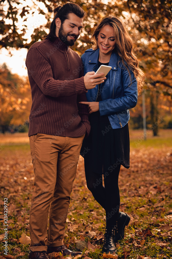 Fototapeta premium Young couple using cellphone in autumn colored park.