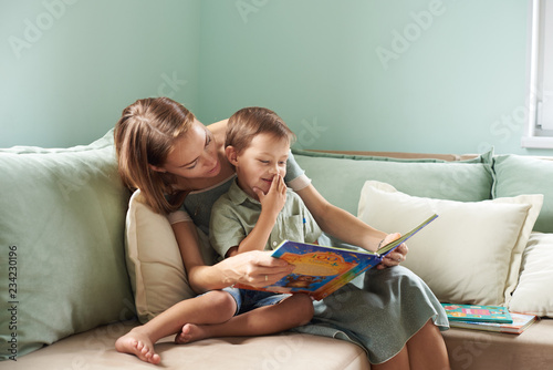 Young mother, read a book to her child, boy in the living room of their home, rays of sun going through the window