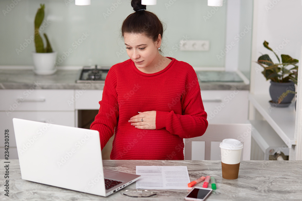 Pregnant woman dressed in red sweater, works from home, keeps hand on belly, looks attentively into laptop, drinks coffee, surrounded with papers, being on maternity leave, poses at kitchen interior Pregnant woman dressed in red sweater, works from home, keeps hand on belly, looks attentively into laptop, drinks coffee, surrounded with papers, being on maternity leave, poses at kitchen interior