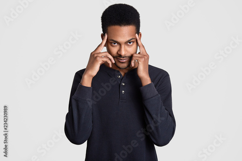 Contemplative dark skinned man keeps both index fingers on temples, thinks about something, tries to concentrate and remember some information, dressed in casual clothes, isolated over white wall