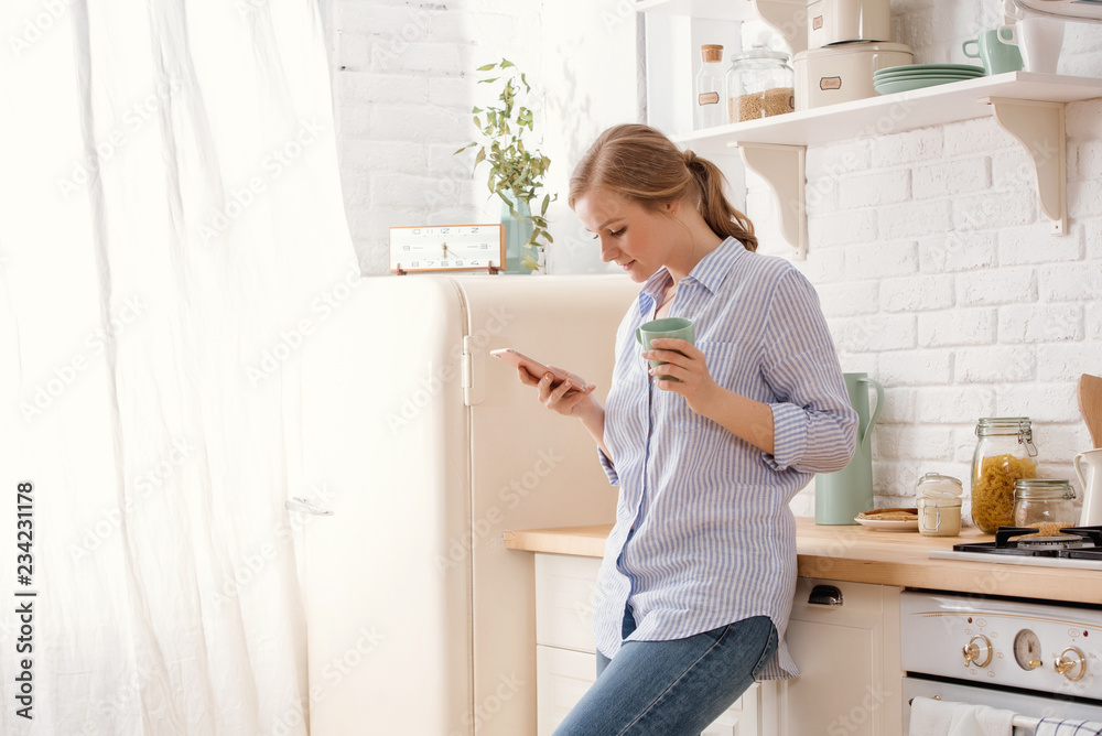 Young woman using smartphone leaning at kitchen table with coffee mug and organizer in a modern home. Smiling woman reading phone message. Brunette happy girl typing a text message. Young woman using smartphone leaning at kitchen table with coffee mug and organizer in a modern home. Smiling woman reading phone message. Brunette happy girl typing a text message.