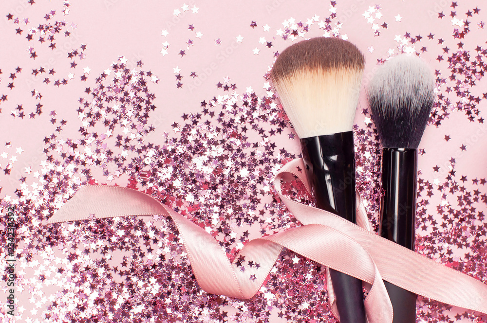 Two different Cosmetic makeup brushes with pink ribbon and holographic  glitter confetti in the form of stars on pink background Flat lay top view  copy space Makeup accessories holiday birthday newyear Stock