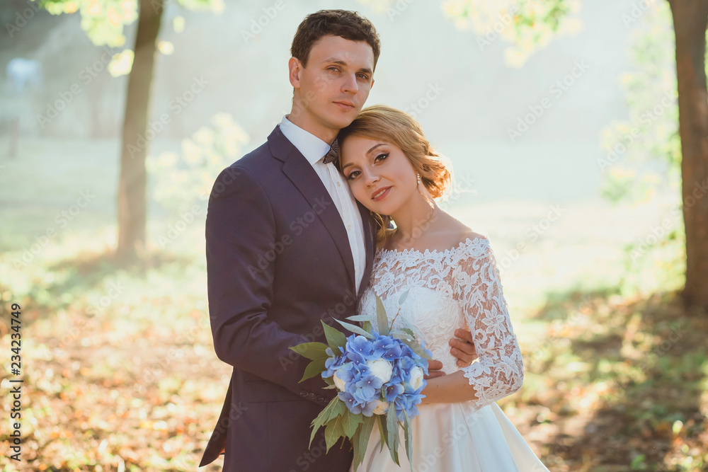 Amazing attractive young couple in wedding day. bride in elegant white long dress and blue bouquet in hand, the groom in a blue fashionable business suit, looking together at the camera