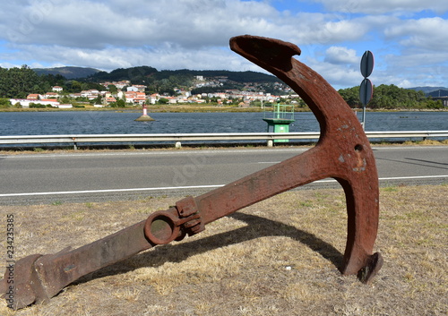 Rusty anchor in a road. River with small lighthouses and coastal village. Pontevedra, Galicia, Spain. Cloudy sky.