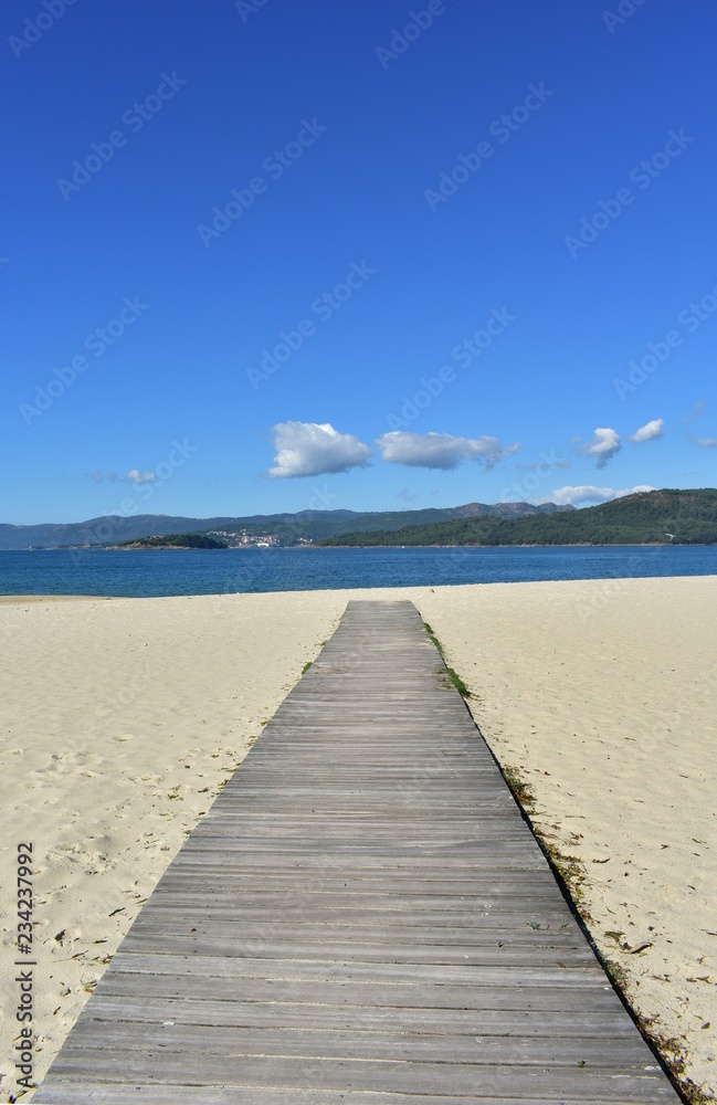 Beach in a bay with bright sand, blue water and wooden boardwalk. Galicia, Spain. Sunny day, blue sky.