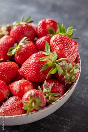 Ripe strawberries with green leaves in a ceramic plate  against a black concrete background. Vitamin healthy berry.