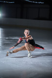 A young ice skater is making spins on the ice rink. She is having a performance in front of the audience.