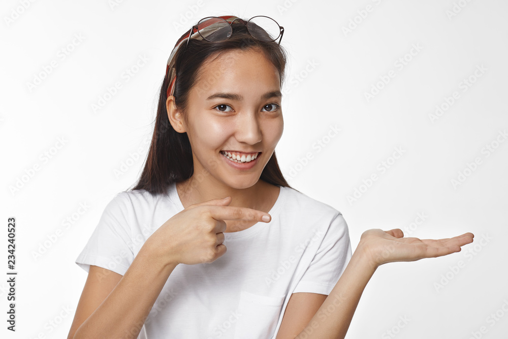 Beautiful positive young Asian female wearing white t-shirt and round glasses on her head keeping hand open as if holding something and pointing fore finger, showing copyspace for your text Beautiful positive young Asian female wearing white t-shirt and round glasses on her head keeping hand open as if holding something and pointing fore finger, showing copyspace for your text