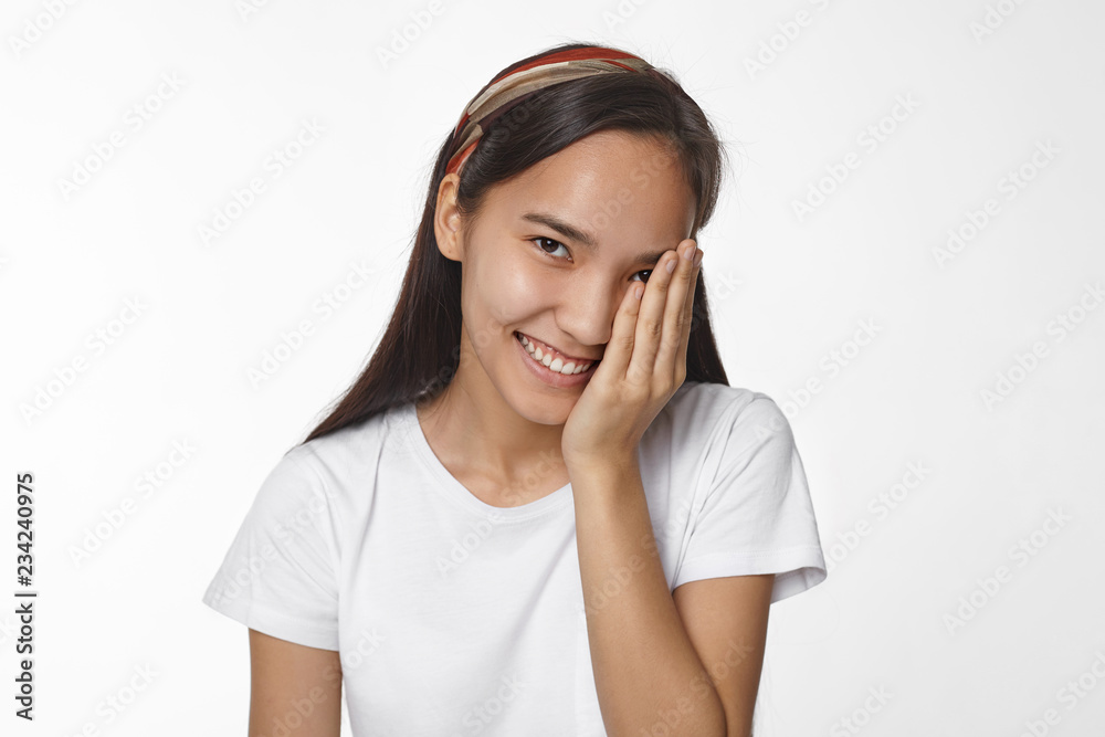 Shyness, youth, people and lifestyle concept. Picture of charming timid young Asian female in white t-shirt looking at camera with cheerful broad smile, covering one eye, being shy and coy Shyness, youth, people and lifestyle concept. Picture of charming timid young Asian female in white t-shirt looking at camera with cheerful broad smile, covering one eye, being shy and coy