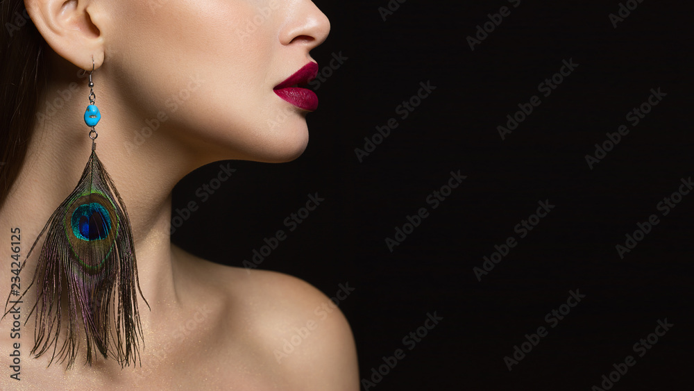 Naklejka premium A close-up portrait of the beauty of a ryunette woman with gorgeous face features, which emphasizes the fatal make-up of red lipstick and fashionable peacock feather earring adds glamor to the image