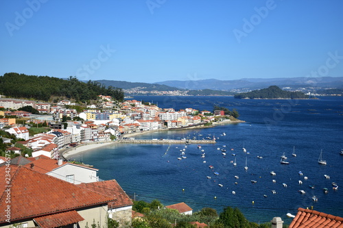 Coastal village in a bay with beach, boats and island with forest. Galicia, Rias Baixas, Spain. Sunny day, blue sky.