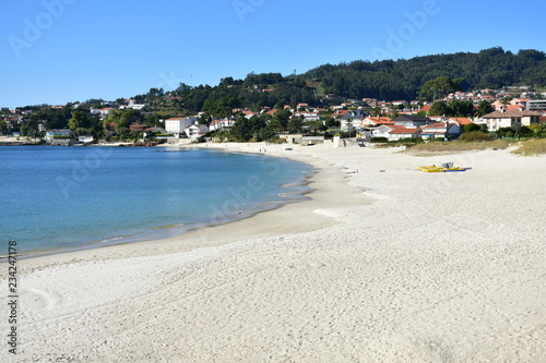 Beach with bright sand and turquoise water. Coastal village, trees and blue sky. Galicia, Rias Baixas, Spain.