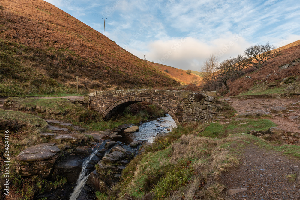 An autumnal waterfall and stone packhorse bridge at Three Shires Head in the Peak District.