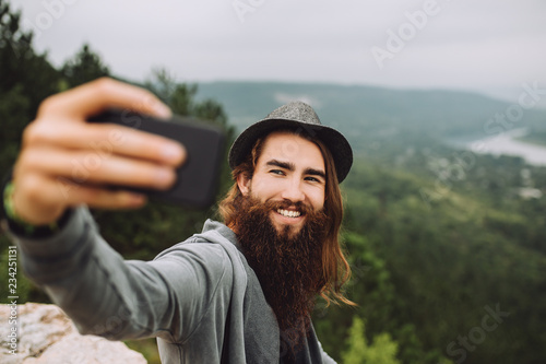 Funny blogger makes selfie on a high cliff in the background of a summer green landscape.The guy holds a fancy smartphone in his hand.A white smile shines on the guy's face.