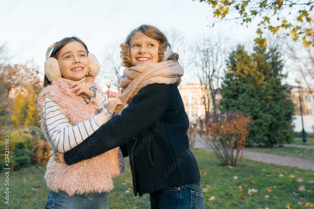 Fototapeta premium Outdoor portrait of two little girls best friends, smiling girls hugging each other watching the sunset, sunny autumn winter park, golden hour